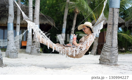 A young woman relaxes in a hammock on a tropical beach working on her laptop while sipping a watermelon shake  130994227