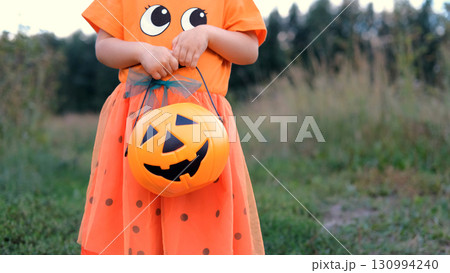 Joyful child girl in a Halloween costume waves while holding a pumpkin shaped basket filled with candy another child dressed as a ghost is seen in the background amid autumn scenery 130994240