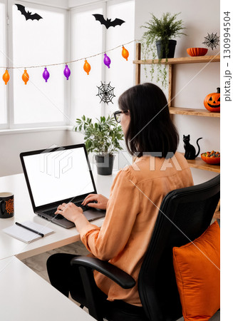 businesswoman working on laptop in room decorated for halloween, monitor mockup, rear view 130994504