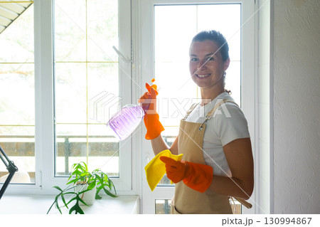 Woman Portrait in rubber gloves and apron manually washes window balcony of house with rag cleaner and mop inside interior. Restoring order and cleanliness in spring, cleaning servise Woman Portrait in rubber gloves and apron manually washes window balcony of house with rag cleaner and mop inside interior. Restoring order and cleanliness in spring, cleaning servise 130994867