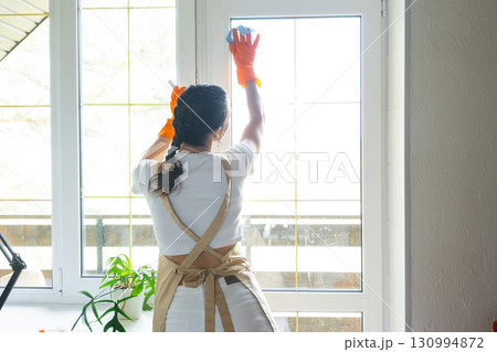 Woman in rubber gloves and apron manually washes window balcony of house with rag cleaner and mop inside interior. Restoring order and cleanliness in spring, cleaning servise 130994872