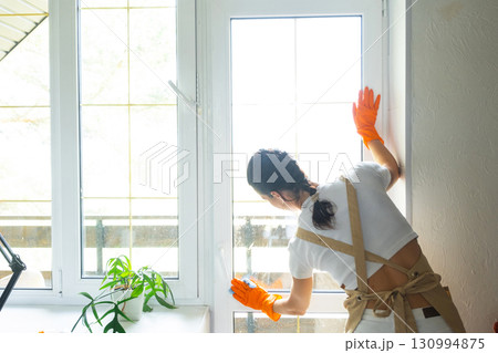Woman in rubber gloves and apron manually washes window balcony of house with rag cleaner and mop inside interior. Restoring order and cleanliness in spring, cleaning servise 130994875