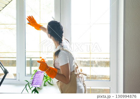 Woman in rubber gloves and apron manually washes window balcony of house with rag cleaner and mop inside interior. Restoring order and cleanliness in spring, cleaning servise 130994876