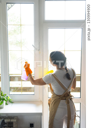 Woman in rubber gloves and apron manually washes window balcony of house with rag cleaner and mop inside interior. Restoring order and cleanliness in spring, cleaning servise 130994900