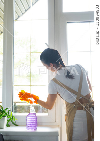 Woman in rubber gloves and apron manually washes window balcony of house with rag cleaner and mop inside interior. Restoring order and cleanliness in spring, cleaning servise 130994903