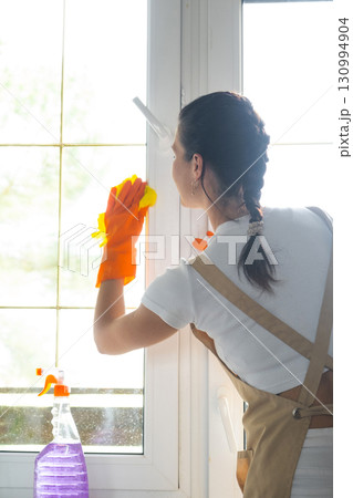 Woman in rubber gloves and apron manually washes window balcony of house with rag cleaner and mop inside interior. Restoring order and cleanliness in spring, cleaning servise 130994904