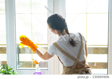 Woman in rubber gloves and apron manually washes window balcony of house with rag cleaner and mop inside interior. Restoring order and cleanliness in spring, cleaning servise 130994906