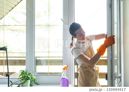 Woman in rubber gloves and apron manually washes window balcony of house with rag cleaner and mop inside interior. Restoring order and cleanliness in spring, cleaning servise Woman in rubber gloves and apron manually washes window balcony of house with rag cleaner and mop inside interior. Restoring order and cleanliness in spring, cleaning servise 130994917