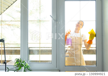 Woman in rubber gloves and apron manually washes window balcony of house with rag cleaner and mop outside behind glass. Restoring order and cleanliness in spring, cleaning servise Woman in rubber gloves and apron manually washes window balcony of house with rag cleaner and mop outside behind glass. Restoring order and cleanliness in spring, cleaning servise 130994921