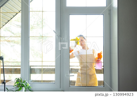 Woman in rubber gloves and apron manually washes window balcony of house with rag cleaner and mop outside behind glass. Restoring order and cleanliness in spring, cleaning servise Woman in rubber gloves and apron manually washes window balcony of house with rag cleaner and mop outside behind glass. Restoring order and cleanliness in spring, cleaning servise 130994923