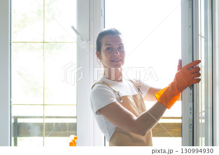 Woman Portrait in rubber gloves and apron manually washes window balcony of house with rag cleaner and mop inside interior. Restoring order and cleanliness in spring, cleaning servise 130994930