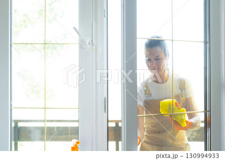 Woman in rubber gloves and apron manually washes window balcony of house with rag cleaner and mop outside behind glass. Restoring order and cleanliness in spring, cleaning servise Woman in rubber gloves and apron manually washes window balcony of house with rag cleaner and mop outside behind glass. Restoring order and cleanliness in spring, cleaning servise 130994933