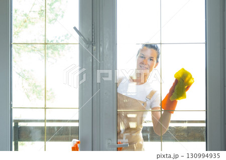 Woman in rubber gloves and apron manually washes window balcony of house with rag cleaner and mop outside behind glass. Restoring order and cleanliness in spring, cleaning servise Woman in rubber gloves and apron manually washes window balcony of house with rag cleaner and mop outside behind glass. Restoring order and cleanliness in spring, cleaning servise 130994935