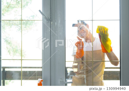 Woman in rubber gloves and apron manually washes window balcony of house with rag cleaner and mop outside behind glass. Restoring order and cleanliness in spring, cleaning servise 130994940