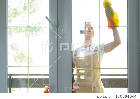 Woman in rubber gloves and apron manually washes window balcony of house with rag cleaner and mop outside behind glass. Restoring order and cleanliness in spring, cleaning servise 130994944
