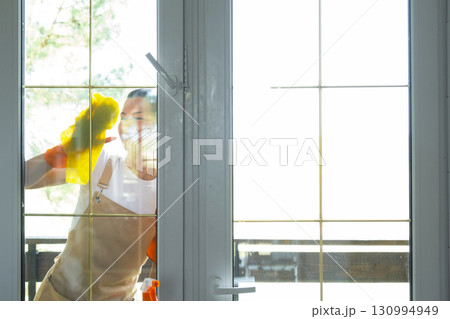 Woman in rubber gloves and apron manually washes window balcony of house with rag cleaner and mop outside behind glass. Restoring order and cleanliness in spring, cleaning servise 130994949