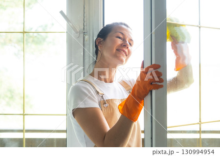 Woman in rubber gloves and apron manually washes window balcony of house with rag cleaner and mop outside behind glass. Restoring order and cleanliness in spring, cleaning servise Woman in rubber gloves and apron manually washes window balcony of house with rag cleaner and mop outside behind glass. Restoring order and cleanliness in spring, cleaning servise 130994954