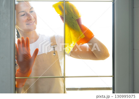 Woman in rubber gloves and apron manually washes window balcony of house with rag cleaner and mop outside behind glass. Restoring order and cleanliness in spring, cleaning servise Woman in rubber gloves and apron manually washes window balcony of house with rag cleaner and mop outside behind glass. Restoring order and cleanliness in spring, cleaning servise 130995075