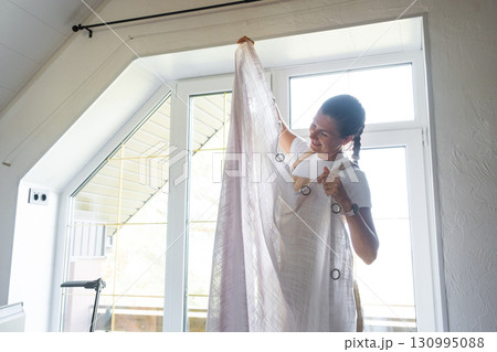 Woman in apron hangs transparent tulle curtains on large attic windows in the house inside the white interior. Spring cleaning, tidying up 130995088