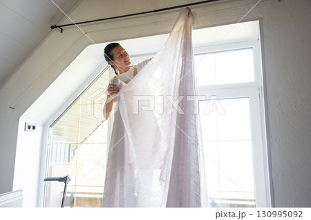Woman in apron hangs transparent tulle curtains on large attic windows in the house inside the white interior. Spring cleaning, tidying up Woman in apron hangs transparent tulle curtains on large attic windows in the house inside the white interior. Spring cleaning, tidying up 130995092