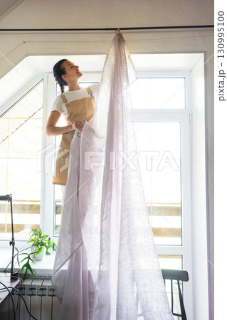 Woman in apron hangs transparent tulle curtains on large attic windows in the house inside the white interior. Spring cleaning, tidying up Woman in apron hangs transparent tulle curtains on large attic windows in the house inside the white interior. Spring cleaning, tidying up 130995100
