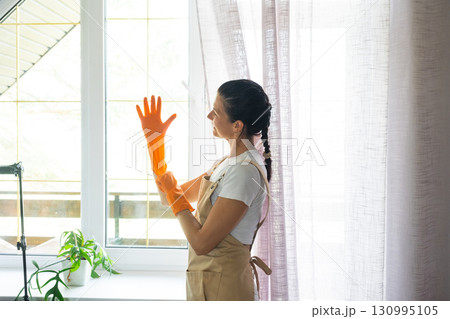 Woman in rubber gloves and apron manually washes window balcony of house with rag cleaner and mop inside interior. Restoring order and cleanliness in spring, cleaning servise Woman in rubber gloves and apron manually washes window balcony of house with rag cleaner and mop inside interior. Restoring order and cleanliness in spring, cleaning servise 130995105