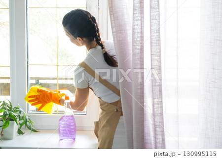 Woman in rubber gloves and apron manually washes window balcony of house with rag cleaner and mop inside interior. Restoring order and cleanliness in spring, cleaning servise 130995115