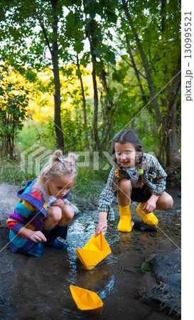 Two girls in yellow and blue boots playing with paper boats in stream Two girls in yellow and blue boots playing with paper boats in stream 130995521