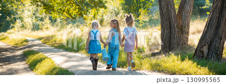 Three little girls holding hands walking with watering can outdoors. Banner 130995565