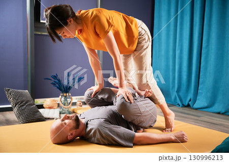 Masseuse making thai yoga massage. Woman therapist doing traditional massage treatment, stretching male patient's body. Bald client lying on mat, enjoying hand massage in calm atmosphere. 130996213