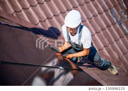 Worker building solar panel system on rooftop of house. Man engineer in helmet installing photovoltaic solar module outdoors. Alternative, green and renewable energy generation concept. 130996220