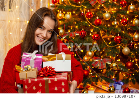 Laughing girl with a pile of presents near Christmas tree. Happy female received many presents at Christmas, showing them to the camera Laughing girl with a pile of presents near Christmas tree. Happy female received many presents at Christmas, showing them to the camera 130997107