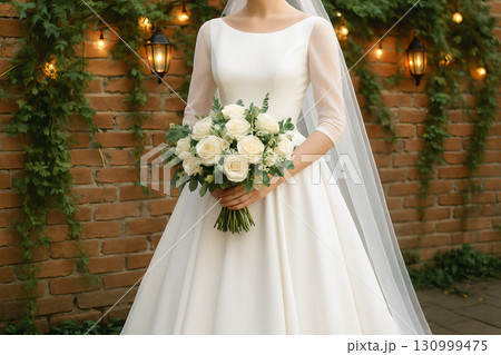 Classic bride holding white rose bouquet next to brick wall decorated with lanterns and greenery. concept of wedding elegance, bridal beauty, timeless romance. 130999475