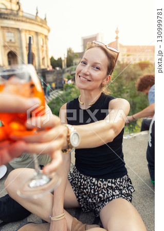 Portrait smiling woman toasts Aperol Spritz historic Berlin architecture museum island at warm evening sunset. Cheerful outdoor moment reflects joyful travel lifestyle relaxed summer city atmosphere 130999781
