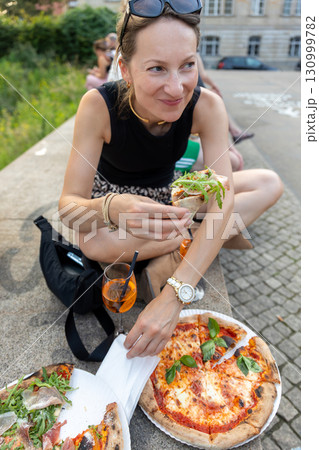 Tourist woman enjoys pizza and Aperol Spritz while sitting outdoors in Berlin during summer day. Relaxed street food atmosphere highlights casual travel lifestyle with authentic culinary experience 130999782