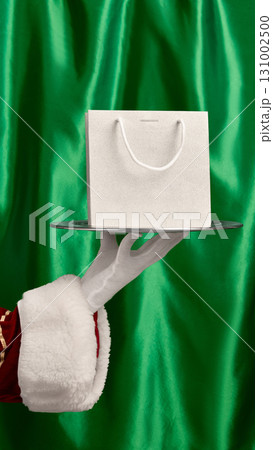 Hand holding gift bag on silver serving tray with green festive curtain. Mockup for ads, text Hand holding gift bag on silver serving tray with green festive curtain. Mockup for ads, text 131002500