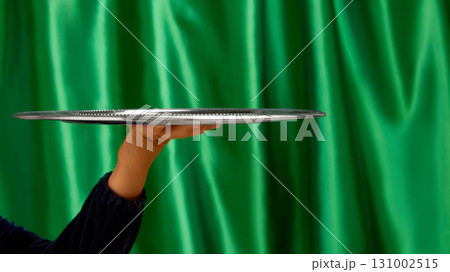 Empty silver serving tray held by bare hand on green curtain background. Negative space for ads 131002515
