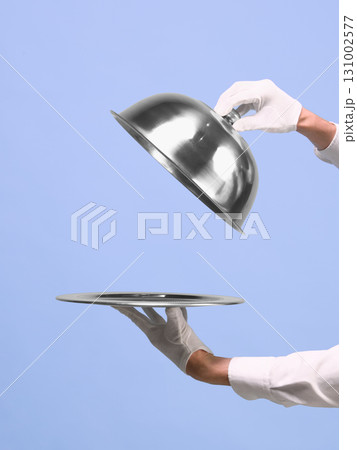 Hands lifting silver cloche from tray on blue background. Negative space for ads 131002577