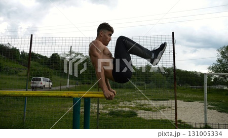 Strong guy training abdominal muscles on parallel bars at sports ground. Young shirtless sportsman exercising at nature. Sporty athlete working out at nature. Concept of active sportive lifestyle Strong guy training abdominal muscles on parallel bars at sports ground. Young shirtless sportsman exercising at nature. Sporty athlete working out at nature. Concept of active sportive lifestyle 131005334