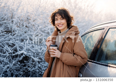 Smiling woman in warm coat holding coffee cup outdoors in frosty winter morning near frozen car Smiling woman in warm coat holding coffee cup outdoors in frosty winter morning near frozen car 131006350