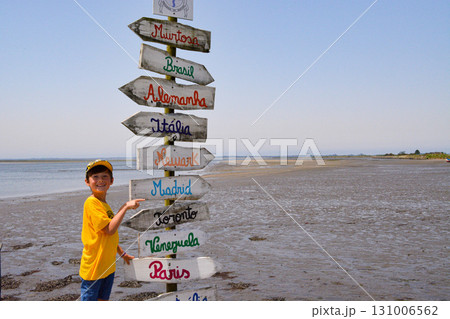 A cheerful, inquisitive toddler boy near colorful wooden signs pointing to all the cities of the world on the seashore. Wanderlust concept. A cheerful, inquisitive toddler boy near colorful wooden signs pointing to all the cities of the world on the seashore. Wanderlust concept. 131006562