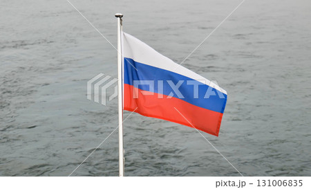 Russian flag waving vigorously, billowing against muted grey waterscape, symbolizing national spirit and patriotic sentiment 131006835
