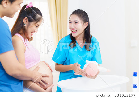 An Asian husband and his pregnant wife are learning how to bathe a newborn baby, guided by a nanny nurse who is demonstrating the bathing process using a baby doll. 131013177