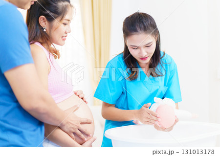 An Asian husband and his pregnant wife are learning how to bathe a newborn baby, guided by a nanny nurse who is demonstrating the bathing process using a baby doll. 131013178