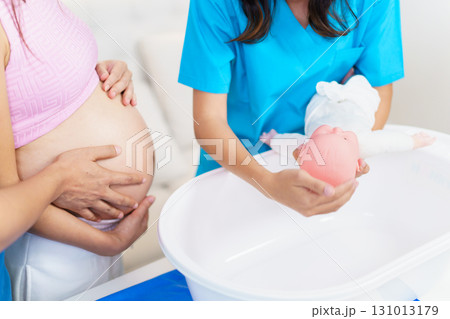 An Asian husband and his pregnant wife are learning how to bathe a newborn baby, guided by a nanny nurse who is demonstrating the bathing process using a baby doll. 131013179