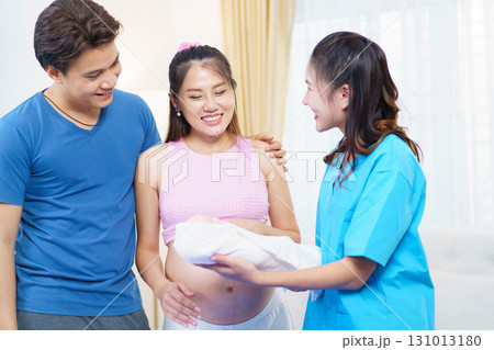 An Asian husband and his pregnant wife are learning how to bathe a newborn baby, guided by a nanny nurse who is demonstrating the bathing process using a baby doll. 131013180