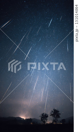 Meteor shower in clear night sky with streaks of light above silhouette trees and distant hills 131014864