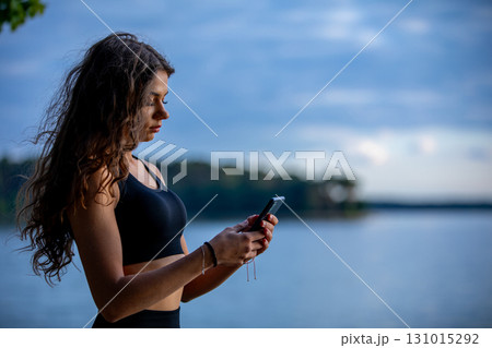 A Young Woman is Engaged with her Smartphone While Relaxing by the Serene Lake at Dusk 131015292