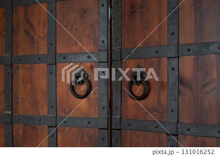 Antique wooden doors with intricate black metal reinforcement. Weathered wooden texture and ornate metalwork on handles showcase craftsmanship and resilience, history and artistry in unique design Antique wooden doors with intricate black metal reinforcement. Weathered wooden texture and ornate metalwork on handles showcase craftsmanship and resilience, history and artistry in unique design 131016252