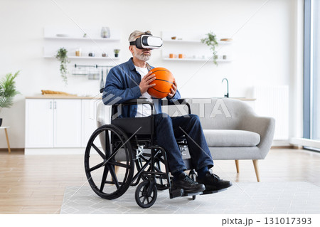 Senior man sitting in wheelchair indoors wears VR headset holding basketball, enjoying technology. Interior setting includes sofa, shelves. Emphasizes virtual reality, accessibility Senior man sitting in wheelchair indoors wears VR headset holding basketball, enjoying technology. Interior setting includes sofa, shelves. Emphasizes virtual reality, accessibility 131017393